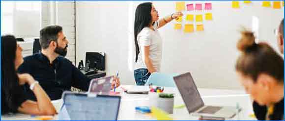 People in a work setting looking at woman pointing at post-its on wall.